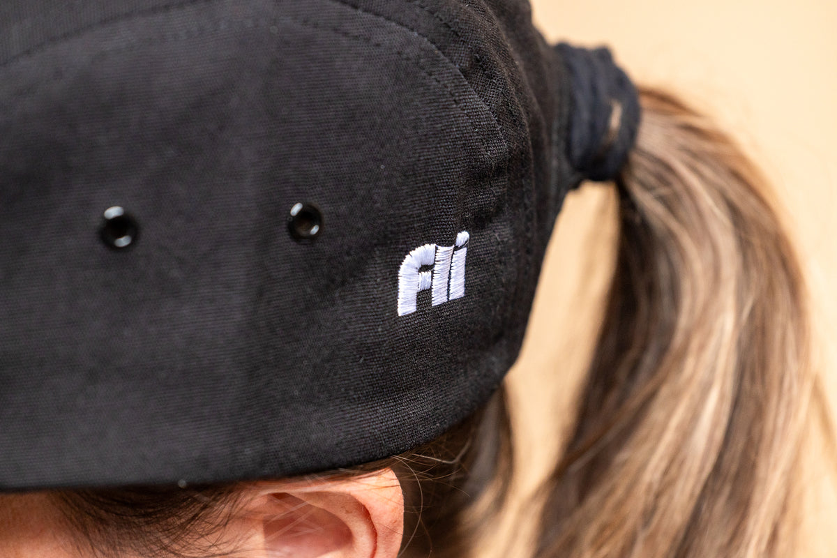 Close-up of a person wearing a black fabric cap oembroidered 'fli' logo, with the hair tied back in a ponytail.