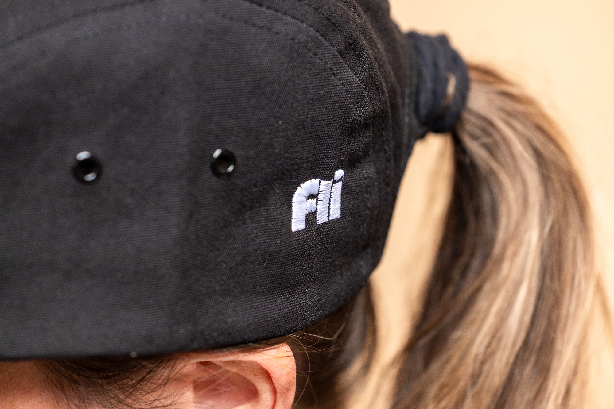 Close-up of a person wearing a black fabric cap oembroidered 'fli' logo, with the hair tied back in a ponytail.