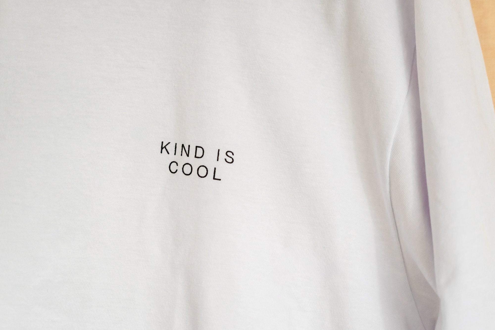 A close-up of a white long-sleeve t-shirt with the words 'KIND IS COOL' printed in black capital letters on the chest.