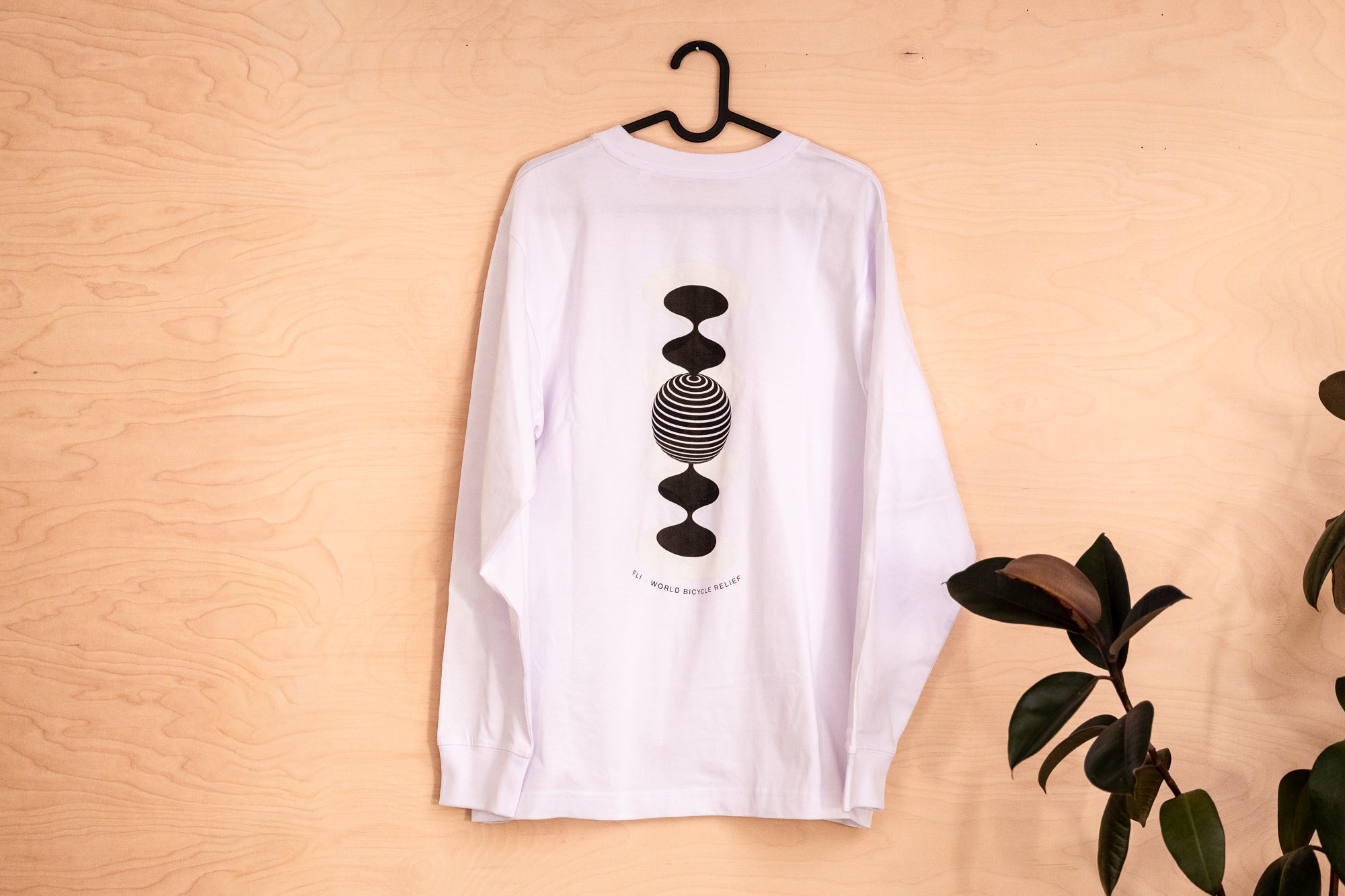 A white long-sleeve t-shirt hanging on a black hanger against a light wood panel background, featuring a black graphic of stacked geometric shapes on the front. With lettering FLi 4 Wordl Bicycle Relief