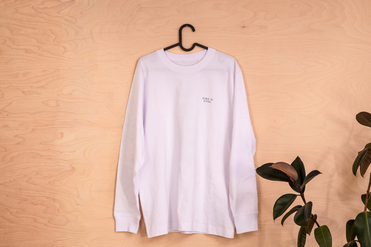 A white long-sleeve crewneck t-shirt with a Kind is cool pring hanging on a black hanger against a light wood panel wall, with a green plant leaf visible on the right side.