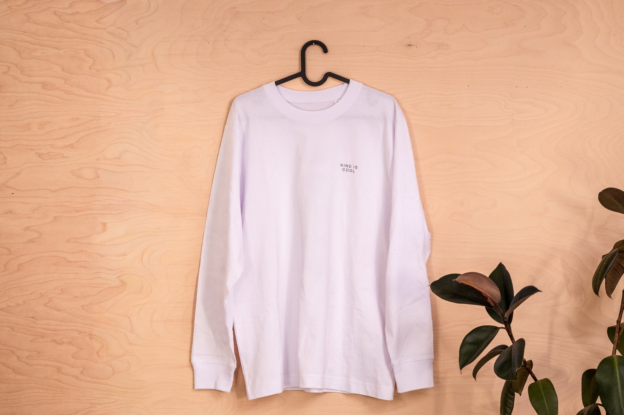 A white long-sleeve crewneck t-shirt with a Kind is cool pring hanging on a black hanger against a light wood panel wall, with a green plant leaf visible on the right side.