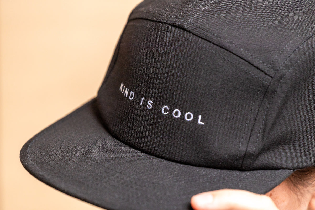 A close-up of a black five-panel cap with 'KIND IS COOL' embroidered in white on the front, worn by a person.