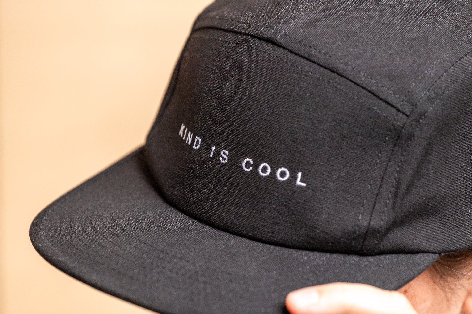 A close-up of a black five-panel cap with 'KIND IS COOL' embroidered in white on the front, worn by a person.