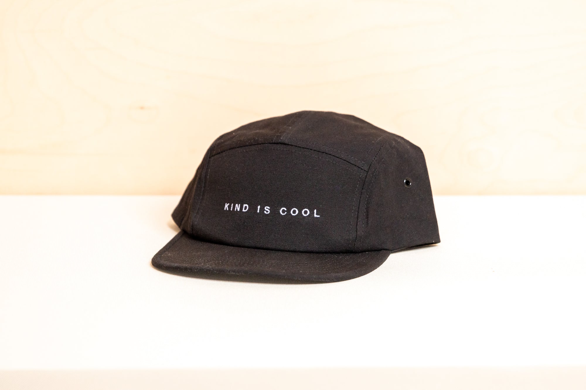 A black five-panel cap with 'KIND IS COOL' embroidered in white on the front, displayed on a white surface.
