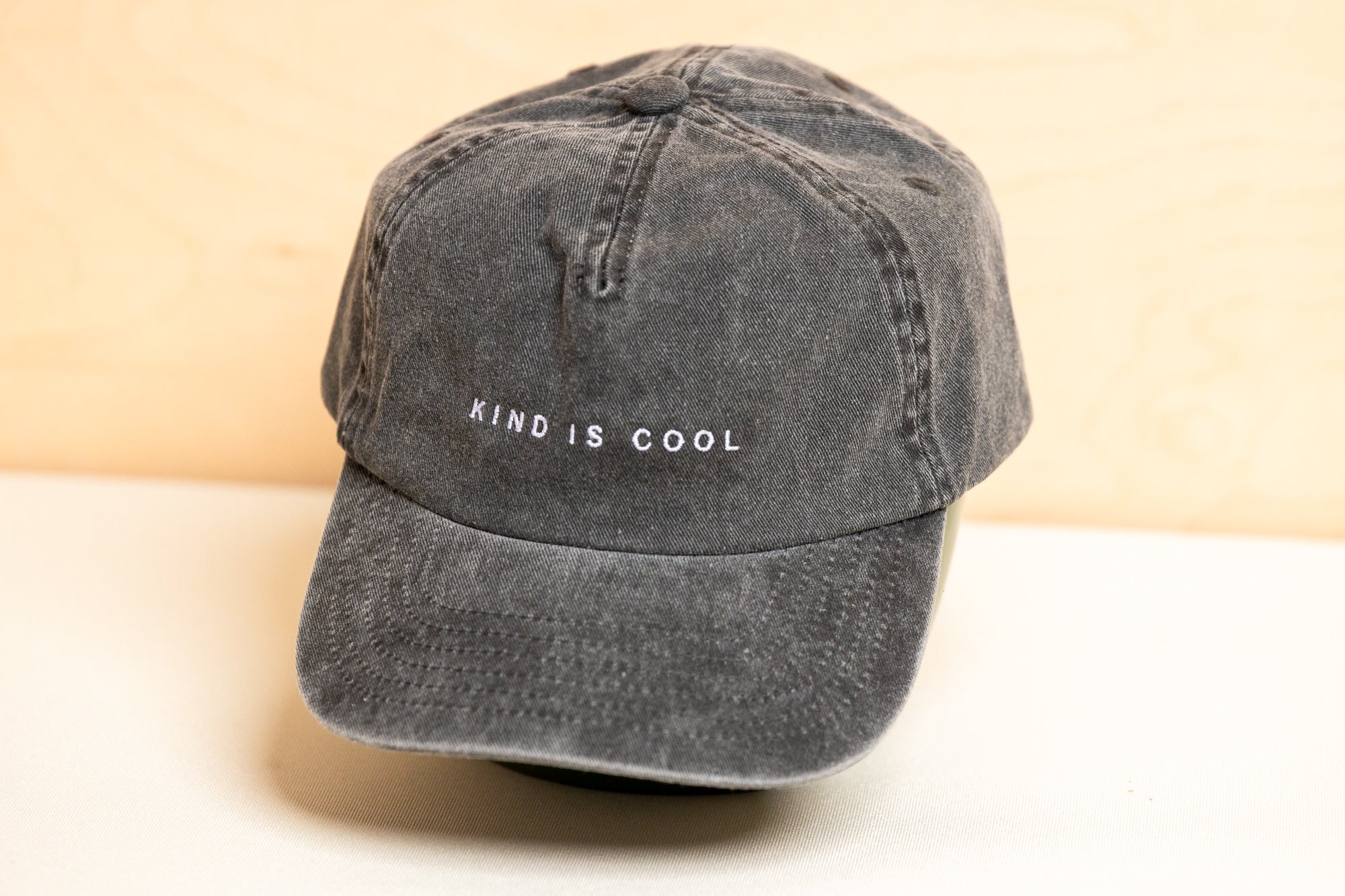 A dark gray washed cotton baseball cap with 'KIND IS COOL' embroidered in white on the front, displayed on a light surface.