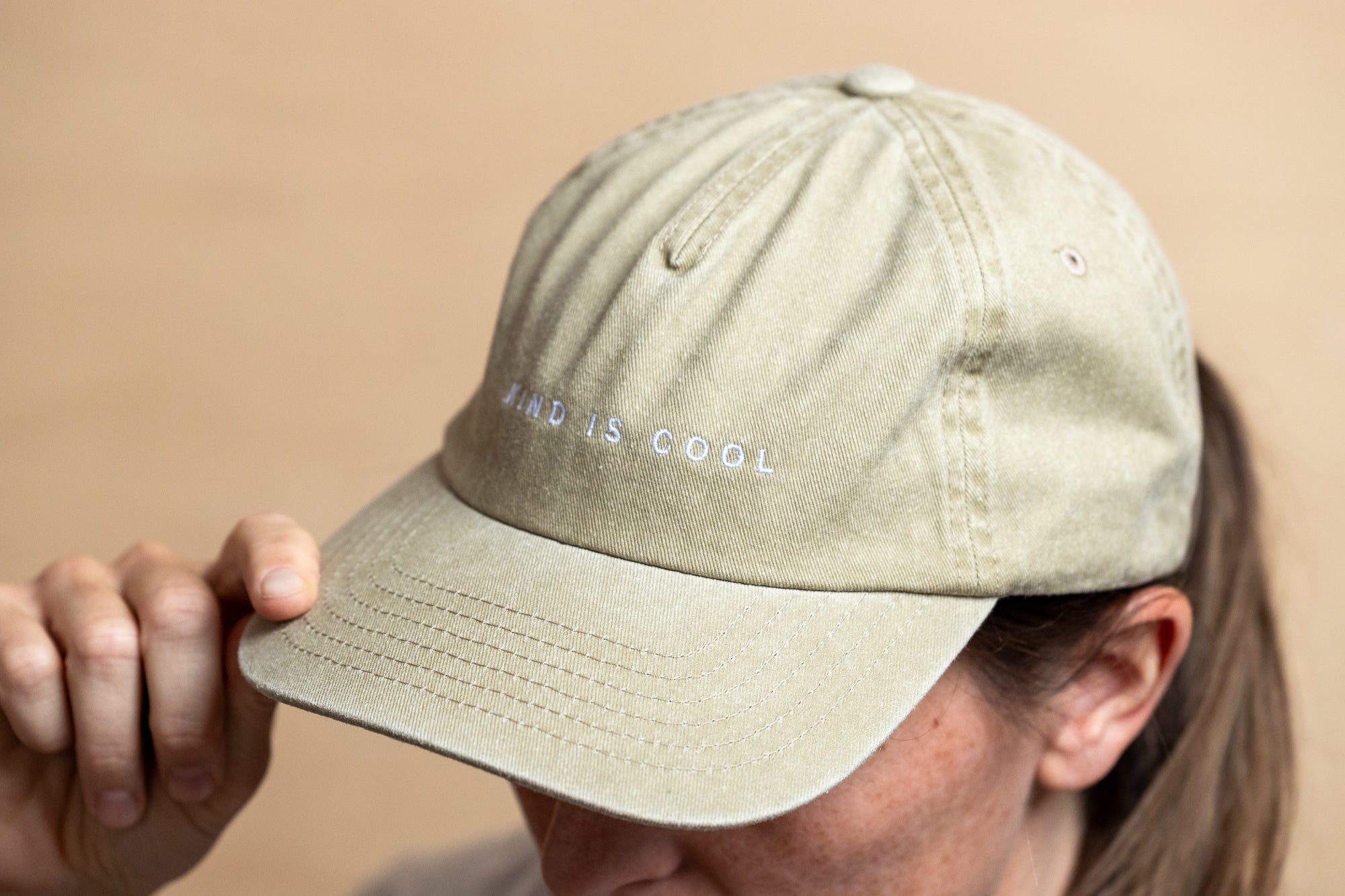 A close-up of a person wearing a light khaki baseball cap with the embroidered text 'KINd IS COOL' on the front, holding the brim with one hand.