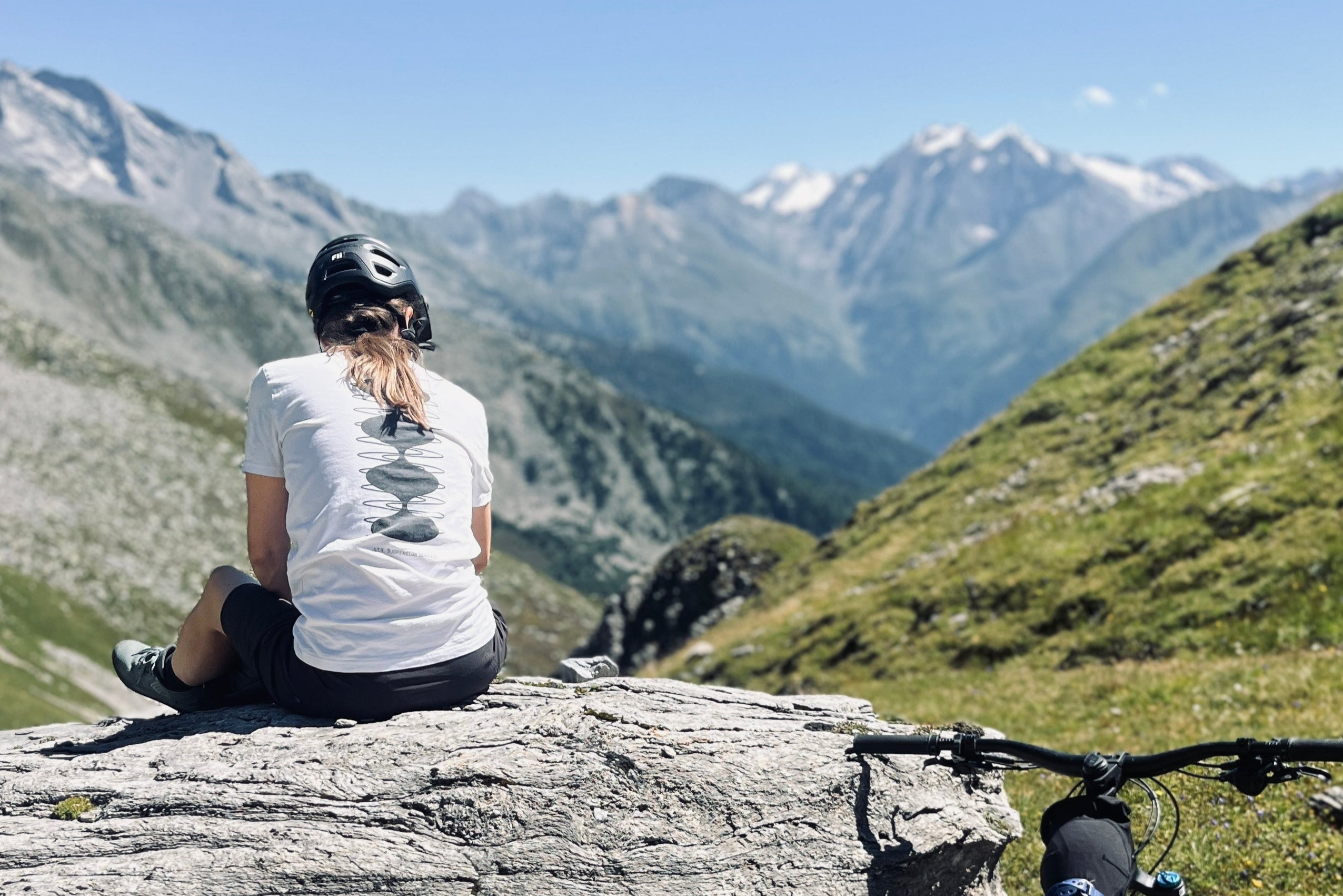 Person sitting on a rock with a mountain bike, overlooking a scenic mountain landscape.