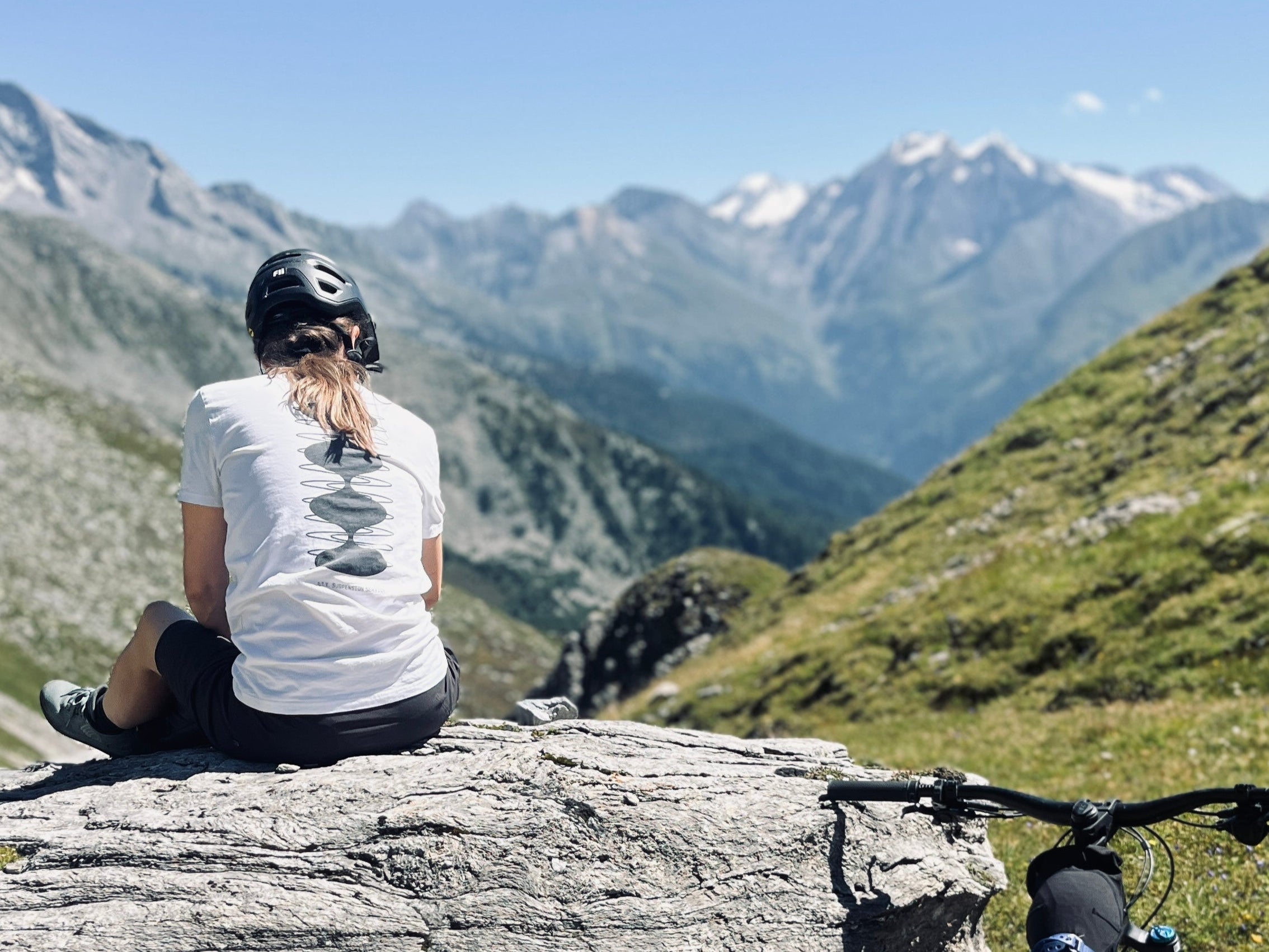 Person sitting on a rock with a mountain bike, overlooking a scenic mountain landscape.