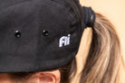Close-up of a person wearing a black fabric cap oembroidered 'fli' logo, with the hair tied back in a ponytail.