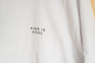 A close-up of a white long-sleeve t-shirt with the words 'KIND IS COOL' printed in black capital letters on the chest.