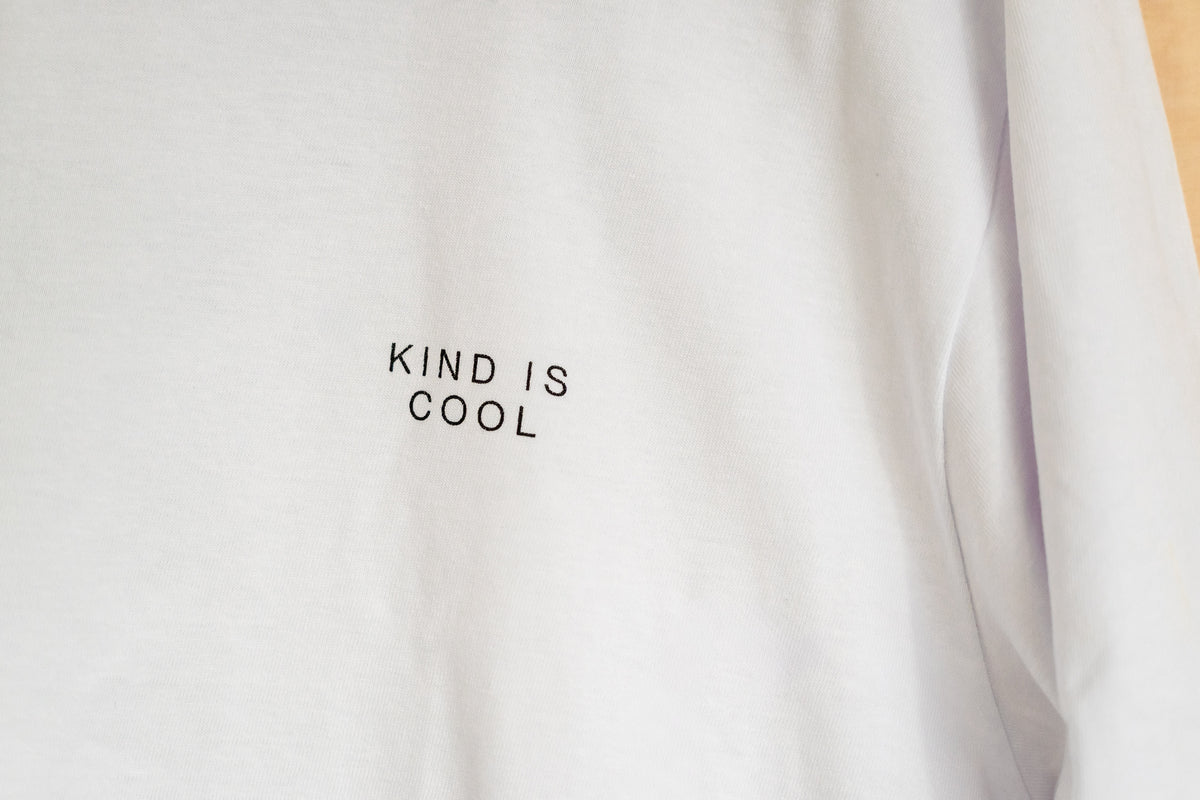 A close-up of a white long-sleeve t-shirt with the words 'KIND IS COOL' printed in black capital letters on the chest.