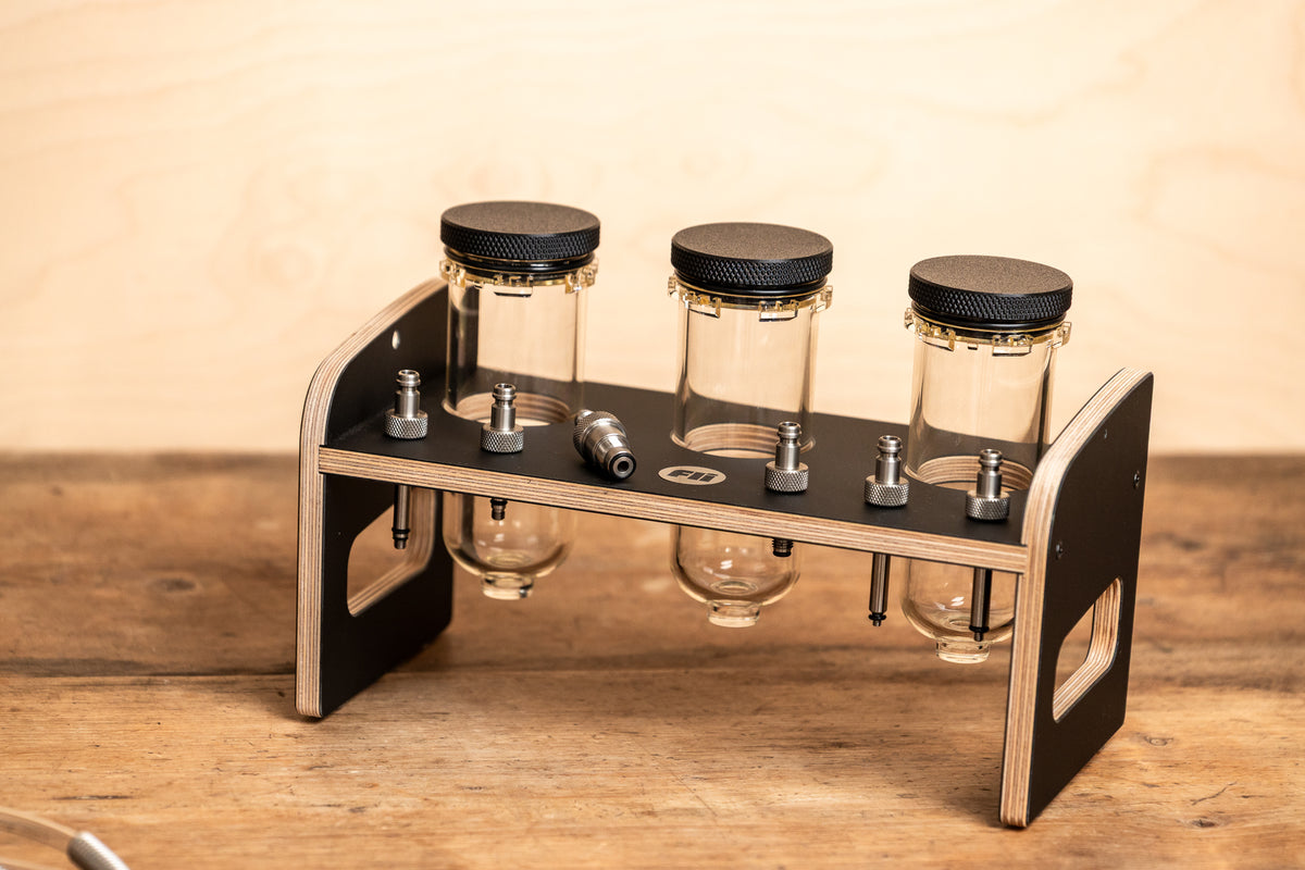 A three-compartment oil tank  and adapter storage for MTB service, made of black and light wood, with three clear tanks, black  lids, and metal adapters, set on a wooden tabletop against a light wood background.