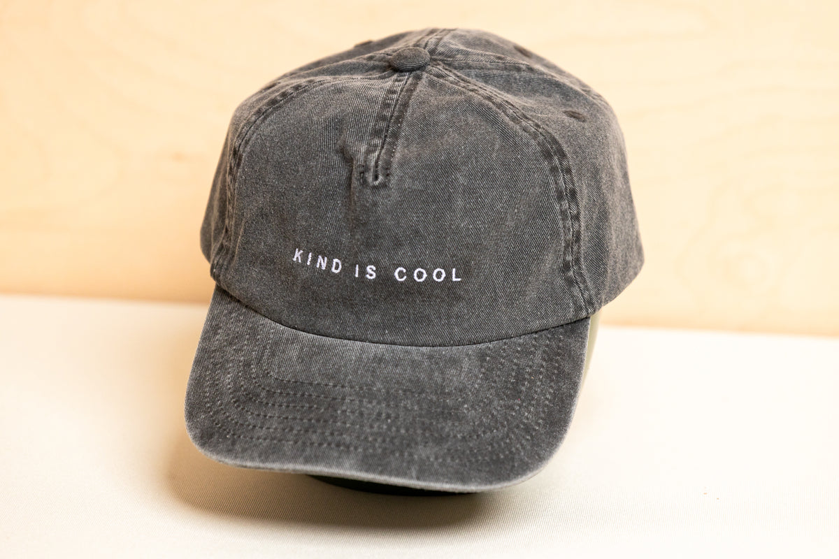 A dark gray washed cotton baseball cap with 'KIND IS COOL' embroidered in white on the front, displayed on a light surface.