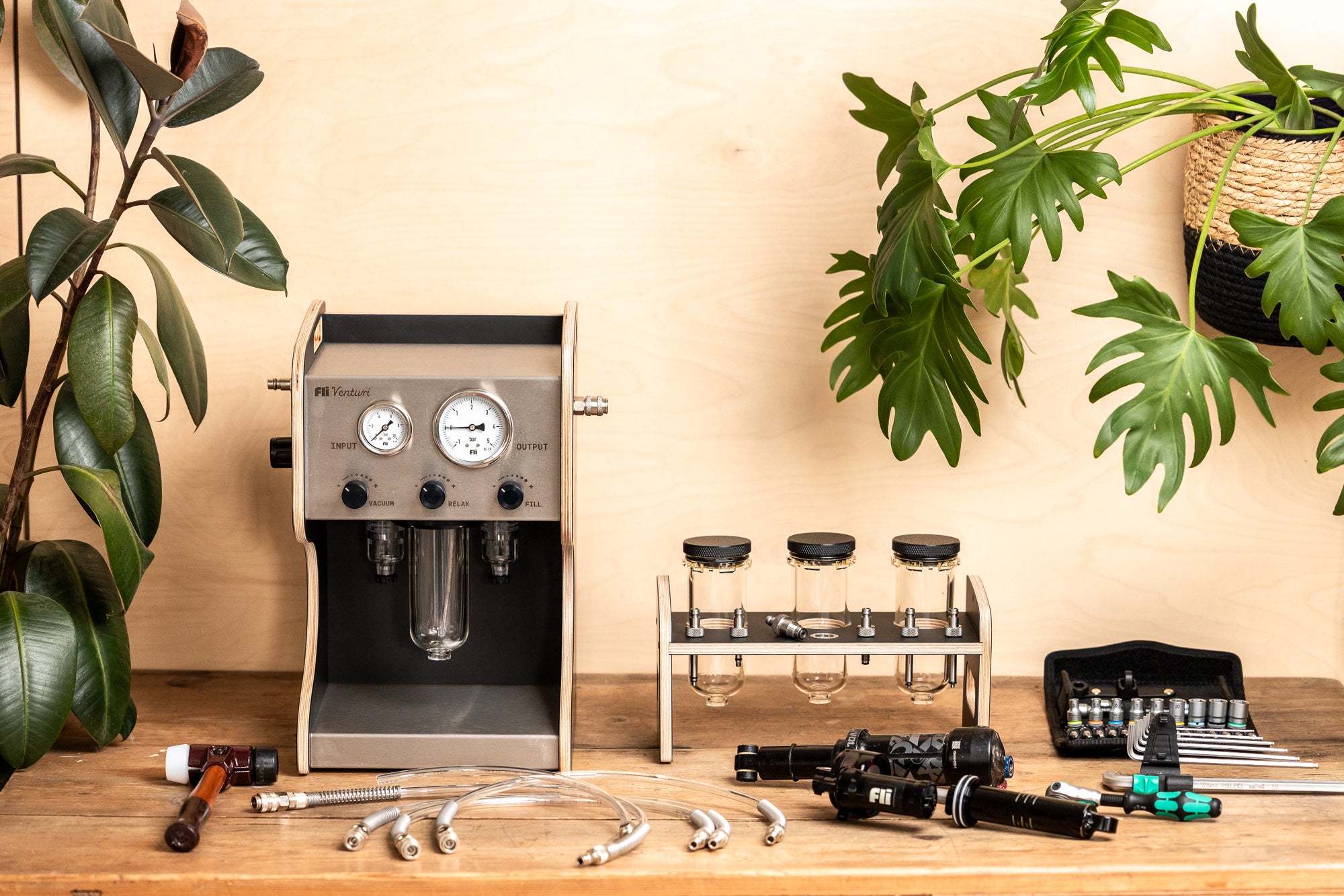 A FLI VENTURI vacuum bleeder resembling an espresso machine, shown with its clear tank, hoses and accessories for suspension service beside damper on a wooden workbench with plants.