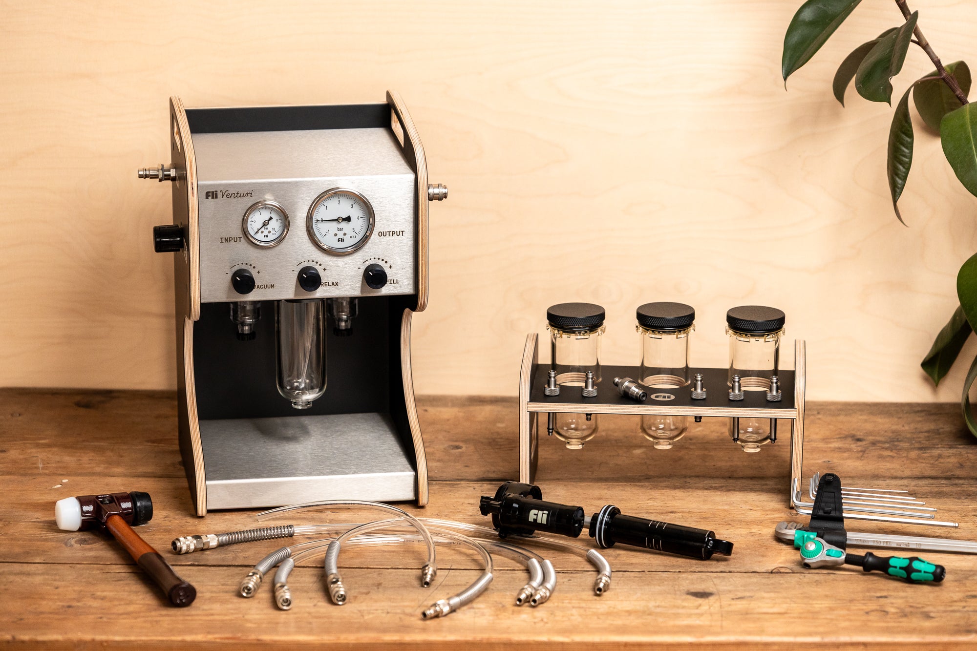 A FLI VENTURI vacuum bleeder resembling an espresso machine, shown with its clear tank, hoses and accessories beside a RockShox damper on a wooden workbench.
