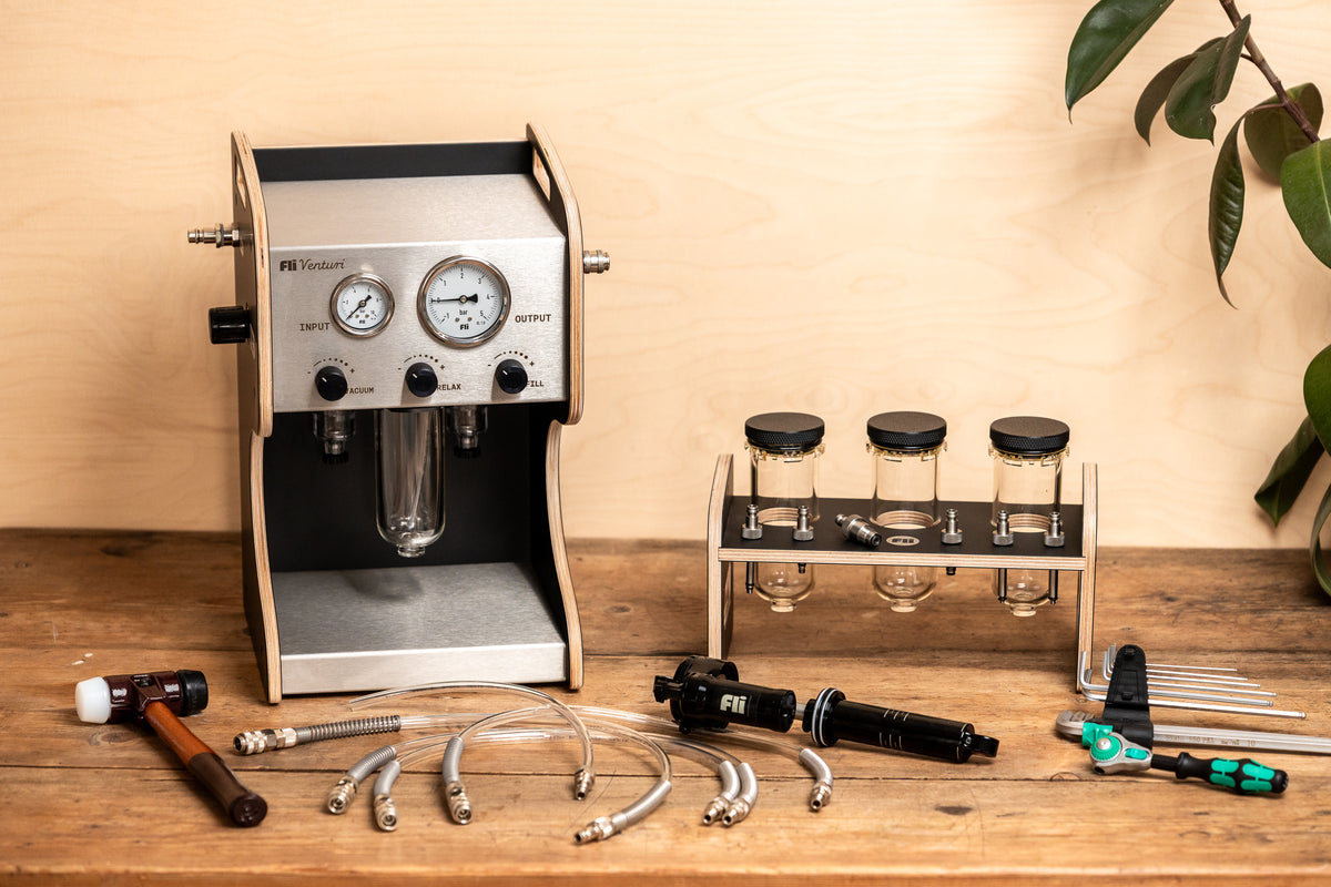 A FLI VENTURI vacuum bleeder resembling an espresso machine, shown with its clear tank, hoses and accessories beside a RockShox damper on a wooden workbench.