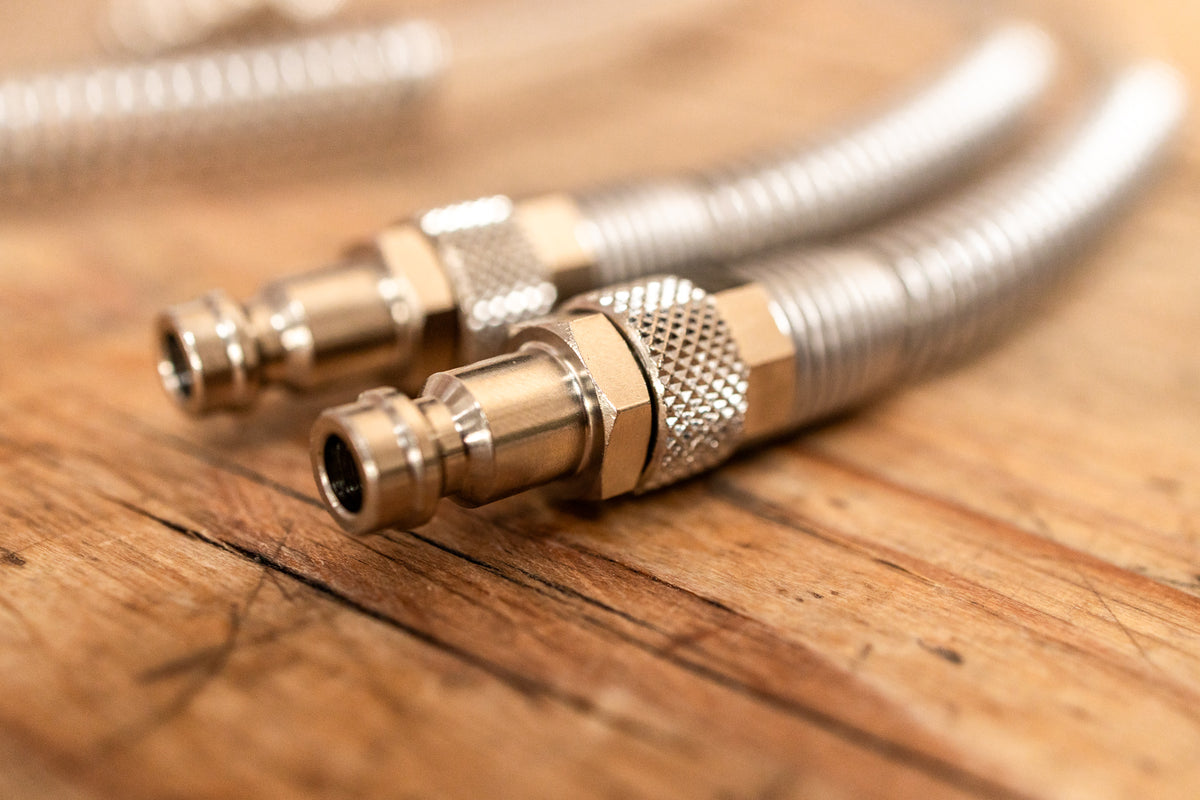 Two metal braided hose connectors with brass fittings resting on a rustic wooden surface.