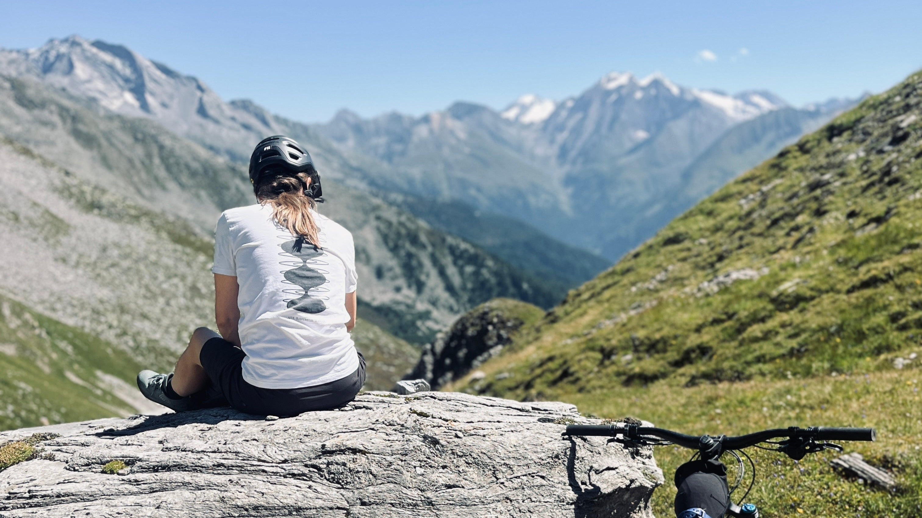 Person sitting on a rock with a mountain bike, overlooking a scenic mountain landscape.